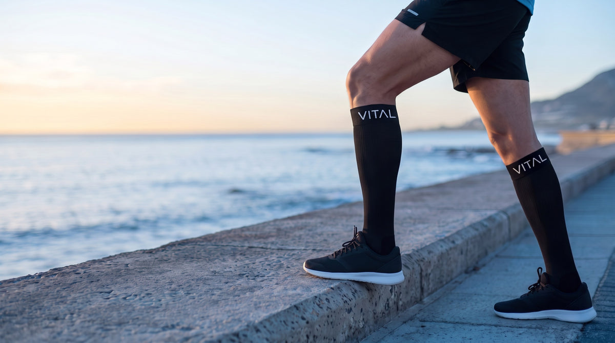 Black knee-high compression socks, black shorts and sneakers, one foot up on a seafront wall