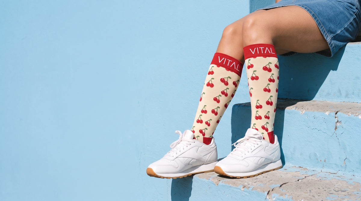 Cherry Pop knee-high compression socks, white leather sneakers and denim shorts, seated on weathered blue steps